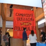 Picture of a banner hanging from a balcony, saying 'Cambridge Sustains Green Colonialism'. A few people are standing in front of the banner.