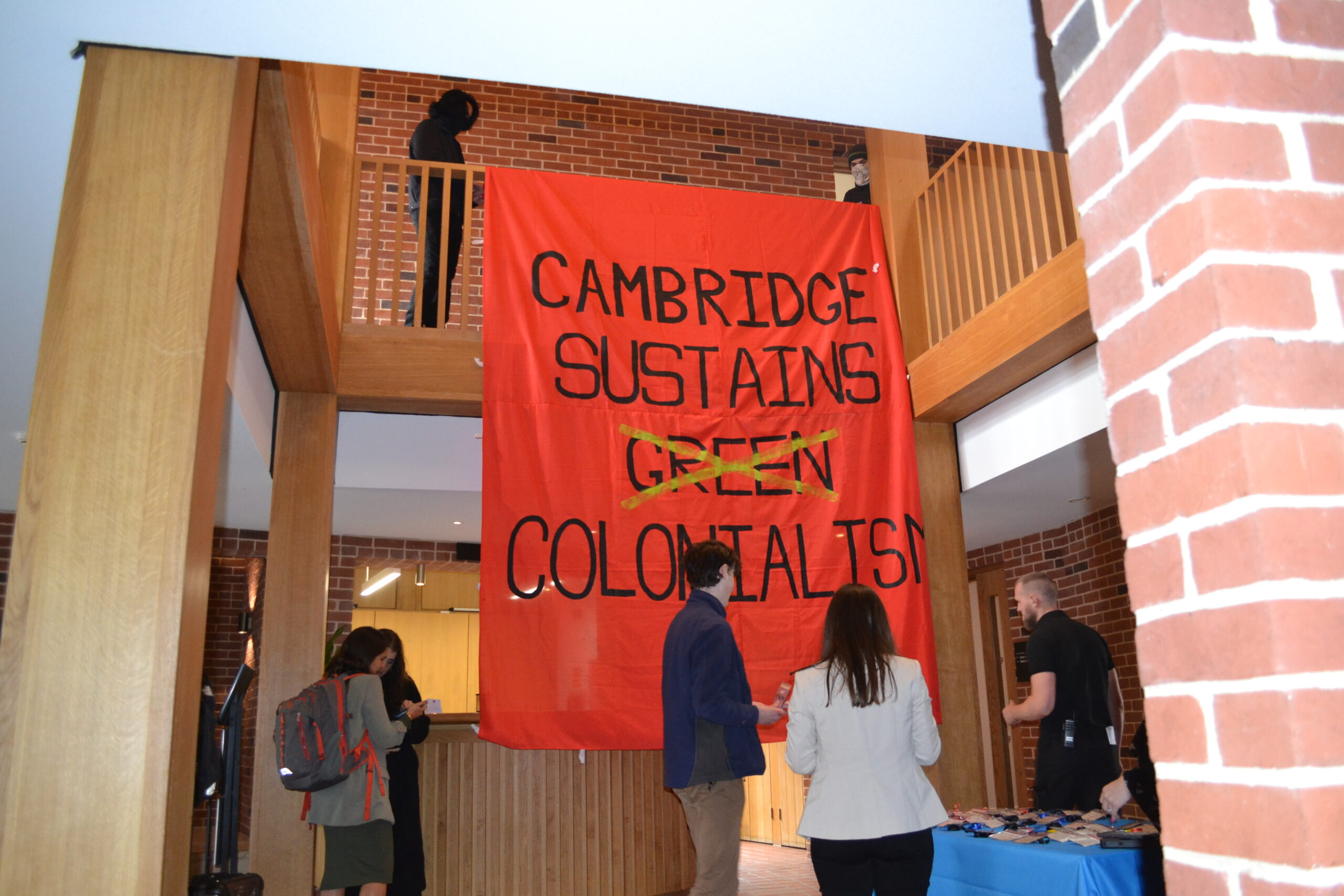 Picture of a banner hanging from a balcony, saying 'Cambridge Sustains Green Colonialism'. A few people are standing in front of the banner.