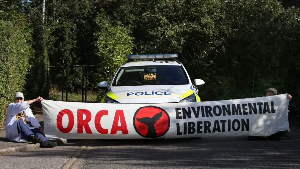 Image of two people holding a banner in front of a police car. One of the 2 has a lab coat. The other one is hidden by the banner. The banner reads "ORCA" in red, then a whale tail, then "environmental liberation" in black. The police car is a standard UK police car, with sirens.
