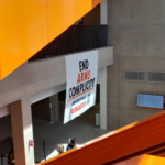 A banner hangs from a balcony. The banner reads "End arms complicity", and there are the logo of Boeing, Rolls-Royce and Bae Systems under the text. The rest of the picture is some concrete architecture elements and orange stairs.