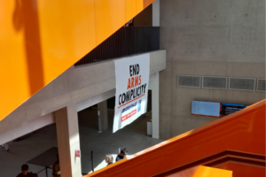 A banner hangs from a balcony. The banner reads "End arms complicity", and there are the logo of Boeing, Rolls-Royce and Bae Systems under the text. The rest of the picture is some concrete architecture elements and orange stairs.