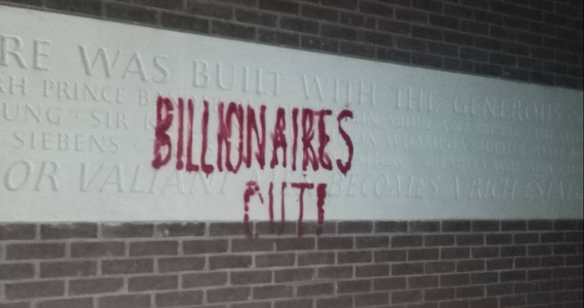A picture of a wall with grafiti-ed on to it "Billionaires out!". The wall features a white stone that thanks the donators to this building, including the Thatcher familly.